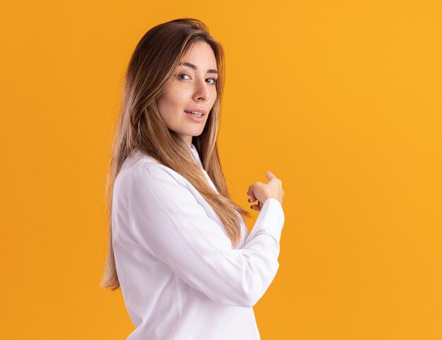 Confident young pretty caucasian girl stands sideways pointing at side isolated on orange wall with copy space