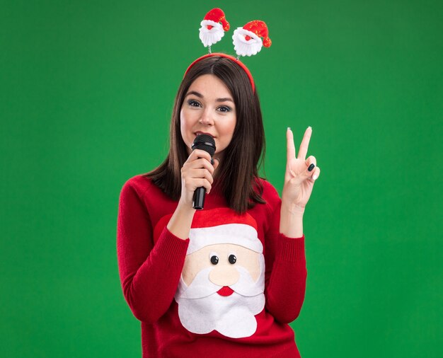 Confident young pretty caucasian girl wearing santa claus sweater and headband talking into microphone  doing peace sign isolated on green wall with copy space