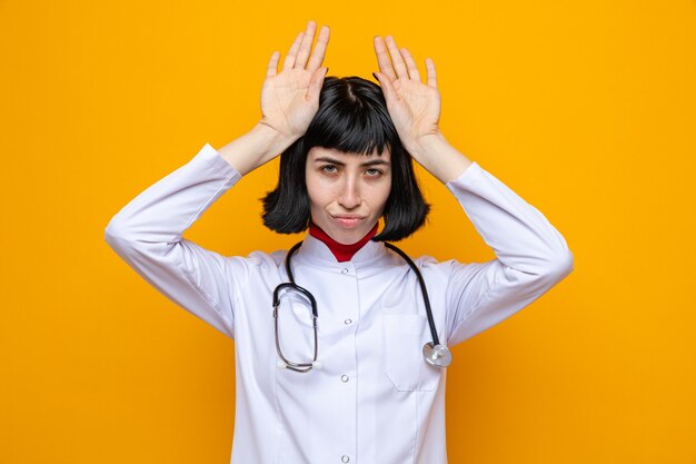 Fiduciosa giovane bella donna caucasica in uniforme da medico con stetoscopio che tiene le mani sulla testa gesticolando le orecchie di coniglio