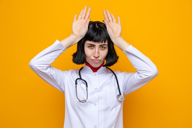 Confident young pretty caucasian woman in doctor uniform with stethoscope keeping hands on her head gesturing rabbit ears 