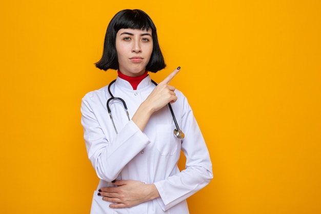 Confident young pretty caucasian woman in doctor uniform with stethoscope pointing at side and putting hand on belly 