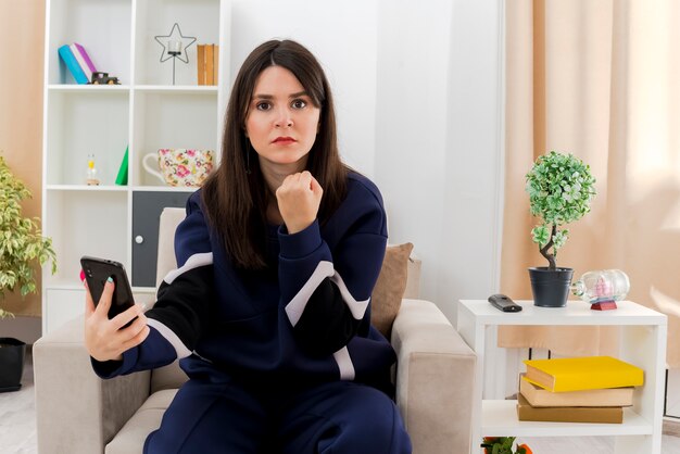 Confident young pretty caucasian woman sitting on armchair in designed living room holding mobile phone clenching fist looking