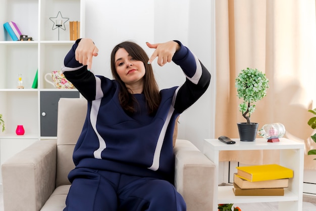 Confident young pretty caucasian woman sitting on armchair in designed living room looking and pointing