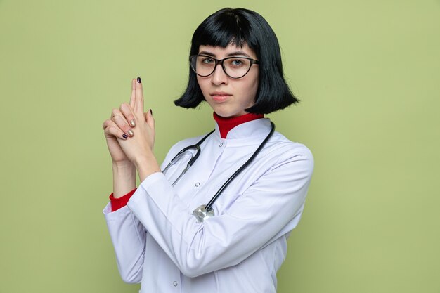 두 손으로 총을 몸짓으로 청진기를 들고 의사 유니폼을 입은 안경을 쓴 자신감 있는 젊은 백인 여성