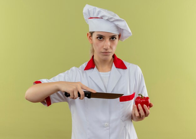 Confident young pretty cook in chef uniform holding pepper and pointing with knife at it  isolated on green wall