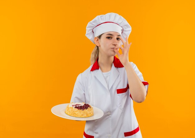 ケーキの皿を持ち、オレンジ色の壁においしいジェスチャーをしているシェフの制服を着た自信のある若いきれいな料理人