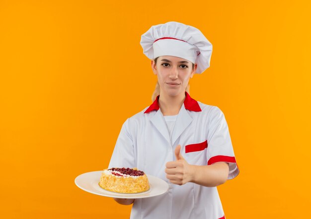 요리사 유니폼 케이크 접시를 들고 엄지 손가락을 보여주는 자신감 젊은 예쁜 요리사는 오렌지 벽에 고립