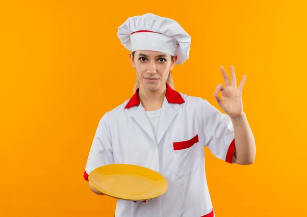 Confident young pretty cook in chef uniform holding plate and doing ok sign isolated on orange wall