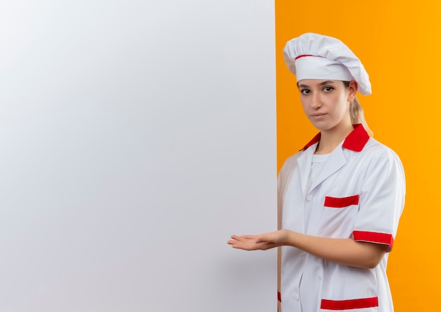 Confident young pretty cook in chef uniform standing near and pointing with hand at white wall isolated on orange wall with copy space