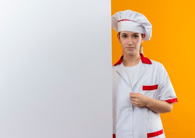 Confident young pretty cook in chef uniform standing behind white wall putting hand on her uniform isolated on orange wall with copy space