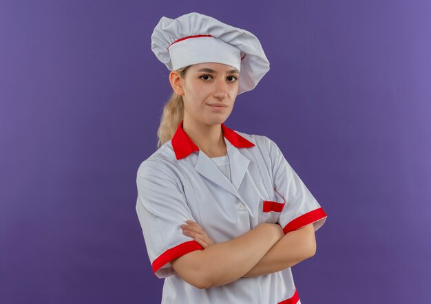 Уверенный молодой симпатичный повар в униформе шеф-повара, стоящий с закрытой позой, выглядит изолированным на фиолетовой стене с копией пространства