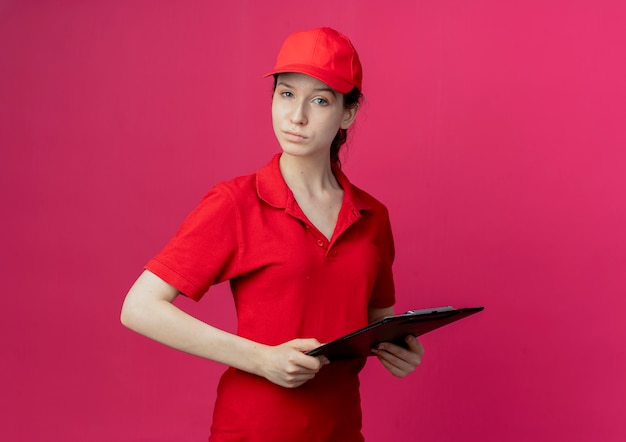 Free photo confident young pretty delivery girl in red uniform and cap holding clipboard looking at camera isolated on crimson background with copy space