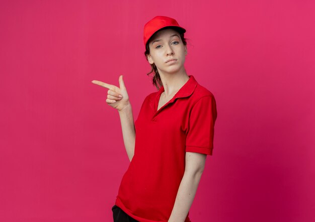 Уверенная молодая симпатичная доставщица в красной форме и кепке, указывающая на сторону, изолированную на малиновом фоне с копией пространства