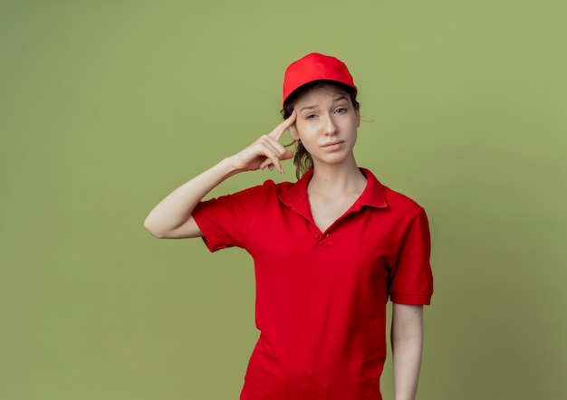 Confident young pretty delivery girl in red uniform and cap putting finger on temple isolated on olive green background with copy space