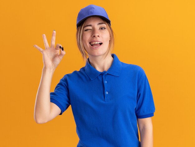 Confident young pretty delivery girl in uniform blinks eye and gestures ok hand sign on orange