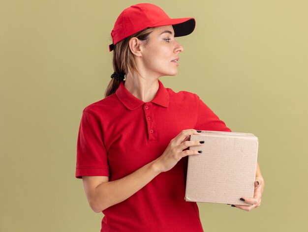 Confident young pretty delivery girl in uniform holds cardbox and looks at side on olive green