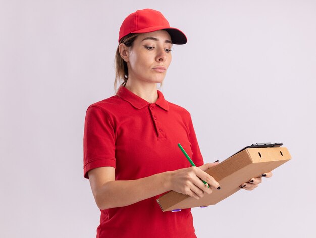 Fiduciosa giovane bella ragazza di consegna in uniforme tiene e guarda negli appunti sulla scatola della pizza su bianco