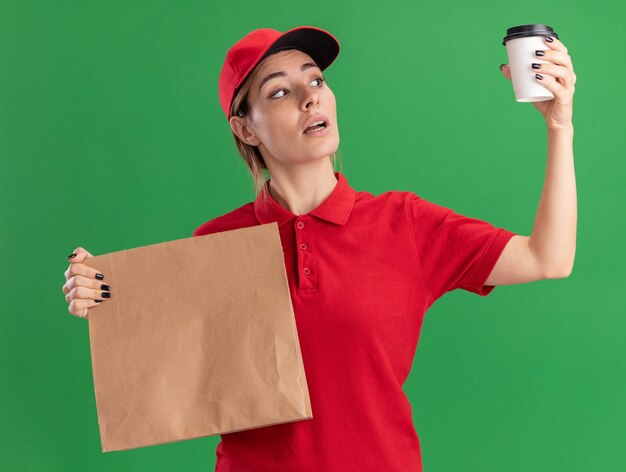 confident young pretty delivery girl in uniform holds paper package and looks at paper cup on green