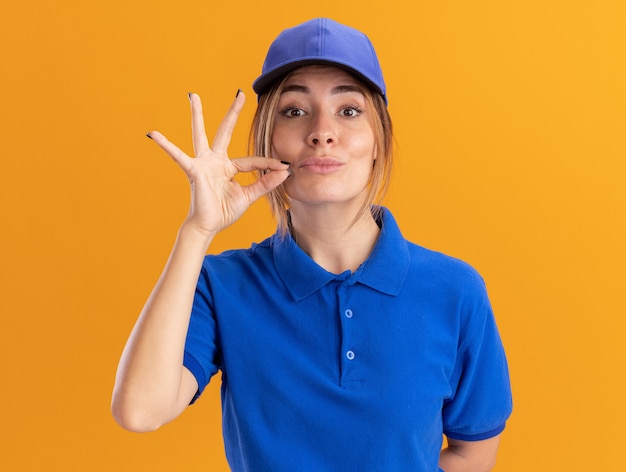 Confident young pretty delivery girl in uniform pretends to zip mouth on orange