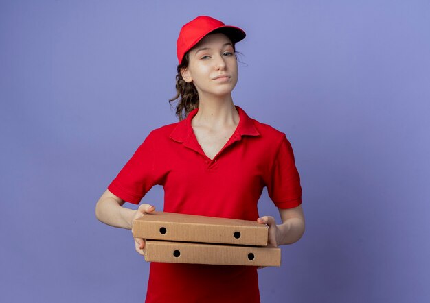 Confident young pretty delivery girl wearing red uniform and cap holding pizza packages isolated on purple background with copy space