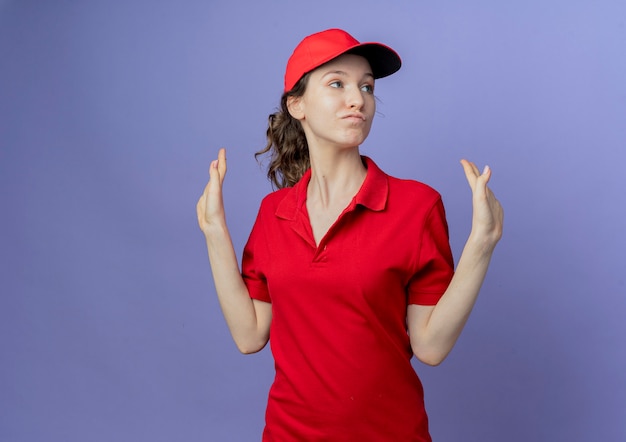 Уверенная молодая симпатичная доставщица в красной форме и кепке, смотрящая в сторону, показывая пустые руки, изолированные на фиолетовом фоне с копией пространства