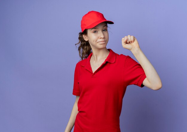 赤い制服とキャップを身に着けている自信を持って若いかわいい配達の女の子は、コピースペースで紫色の背景に分離された上げられた握りこぶしのジェスチャーを示しています