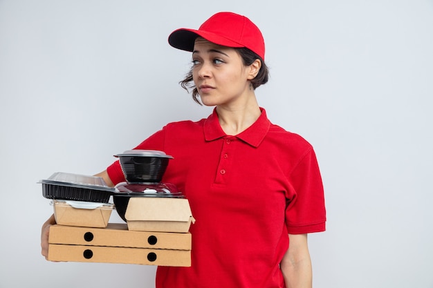 Confident young pretty delivery woman holding food containers and packaging on pizza boxes looking at side 