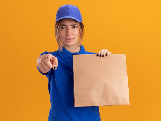 Confident young pretty delivery woman in uniform holds paper package and points at front isolated on orange wall