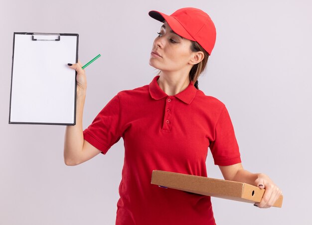 Confident young pretty delivery woman in uniform holds pizza box and looks at clipboard isolated on white wall