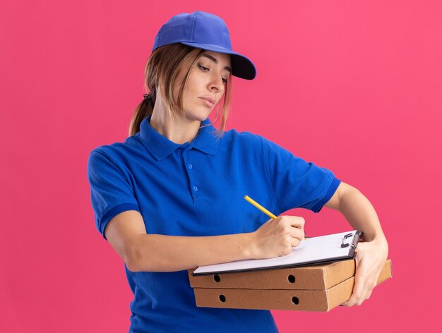 Уверенная молодая красивая женщина-доставщик в униформе держит коробки для пиццы и пишет в буфер обмена ручкой, изолированной на розовой стене