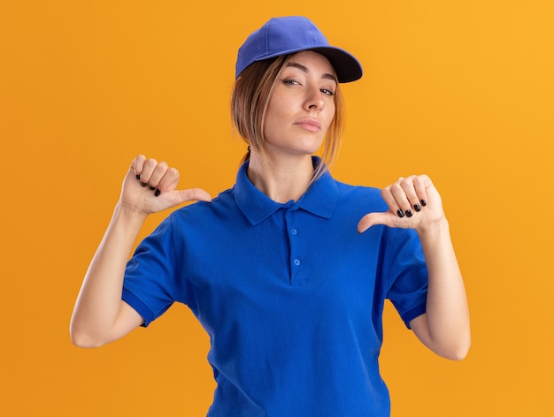 Confident young pretty delivery woman in uniform points at himself with two hands isolated on orange wall