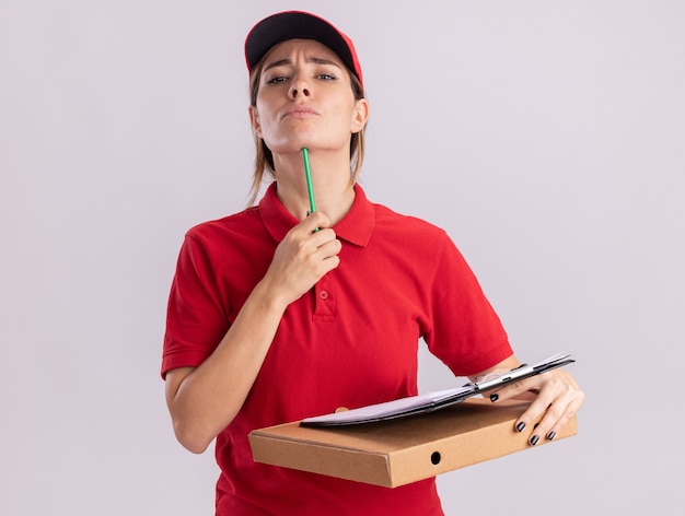 Confident young pretty delivery woman in uniform puts pencil on chin and holds clipboard on pizza box isolated on white wall