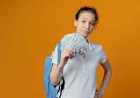 Бесплатное фото Уверенная молодая симпатичная студентка в задней сумке, держащая деньги, кладет руку на талию, изолированную на оранжевом фоне с копией пространства