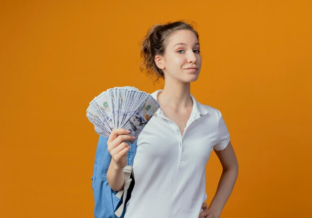 Confident young pretty female student wearing back bag holding money putting hand on waist isolated on orange background with copy space