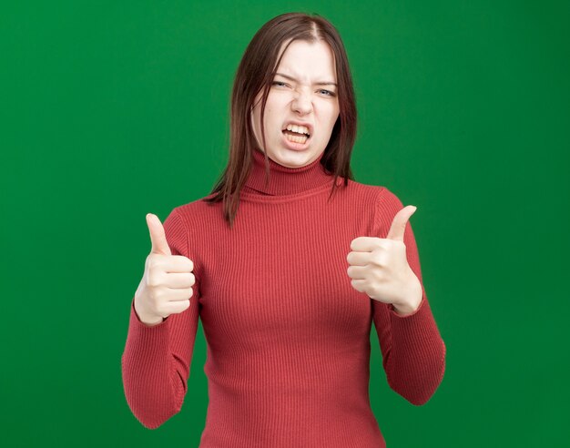 Confident young pretty girl  showing thumbs up isolated on green wall