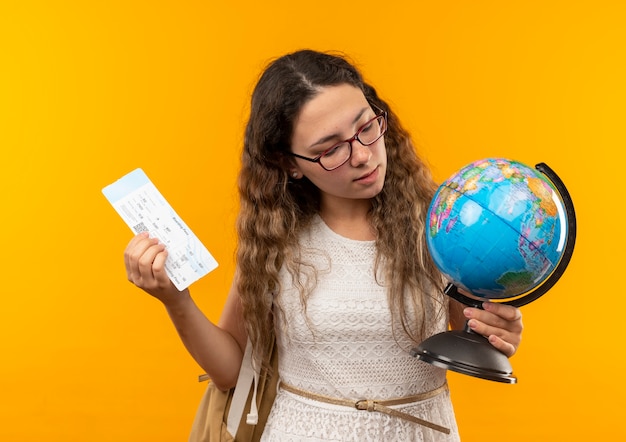 Fiducioso giovane studentessa graziosa con gli occhiali e borsa posteriore in possesso di biglietto aereo e globo guardando globo isolato su giallo