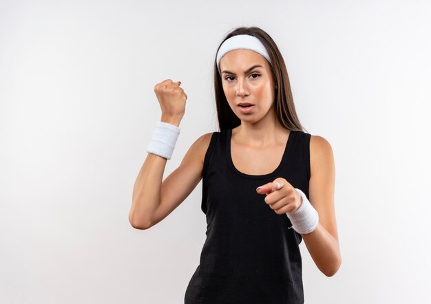 Уверенная молодая симпатичная спортивная девушка с головной повязкой и браслетом, сжимая кулак и указывая на белую стену с копией пространства