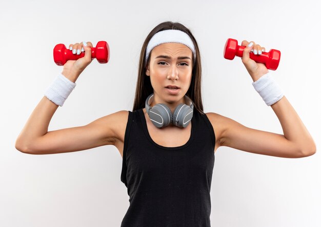 Free photo confident young pretty sporty girl wearing headband and wristband and headphones on neck raising dumbbells isolated on white wall