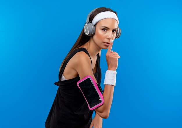 Confident young pretty sporty girl wearing headband and wristband and headphones with phone armband standing in profile view gesturing silence isolated on blue wall