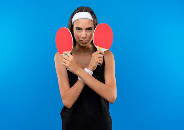 Confident young pretty sporty girl wearing headband and wristband holding ping pong rackets isolated on blue wall with copy space