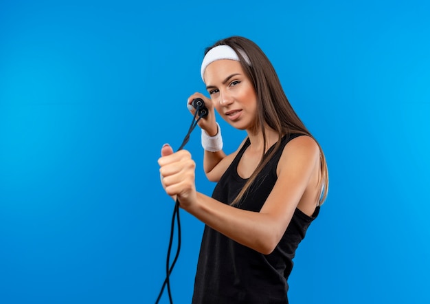 Confident young pretty sporty girl wearing headband and wristband holding and stretching out jumping rope  isolated on blue wall with copy space