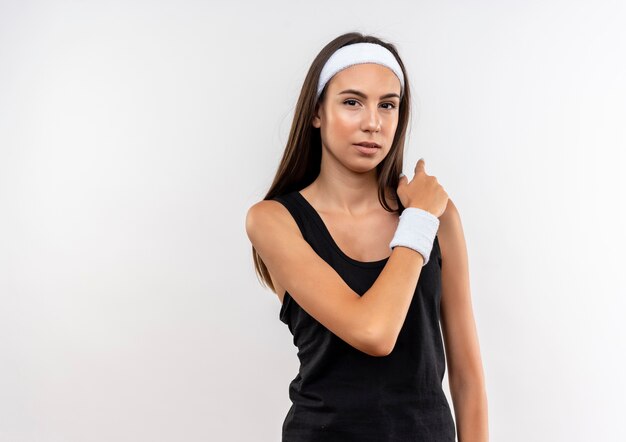 Confident young pretty sporty girl wearing headband and wristband pointing behind on white wall with copy space