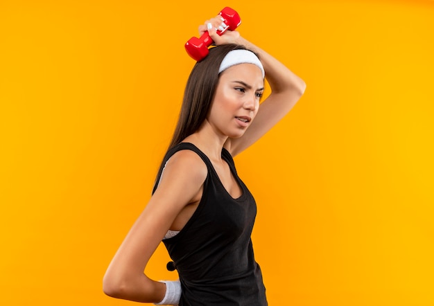 Confident young pretty sporty girl wearing headband and wristband putting dumbbell on head standing in profile view and looking at side isolated on orange wall