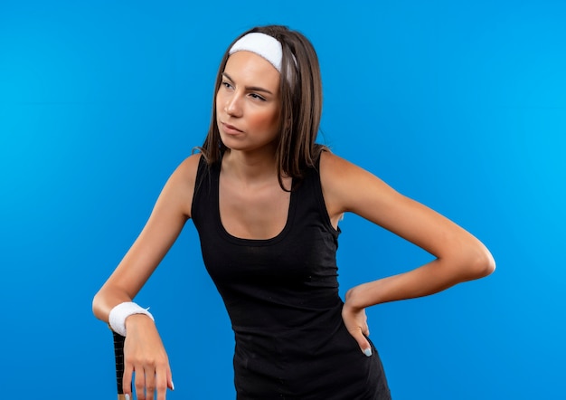 Fiduciosa giovane ragazza abbastanza sportiva che indossa fascia e cinturino che mette le mani sulla mazza da baseball e sulla vita guardando il lato isolato sulla parete blu