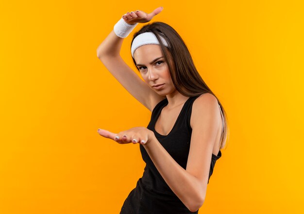 Confident young pretty sporty girl wearing headband and wristband showing size isolated on orange wall