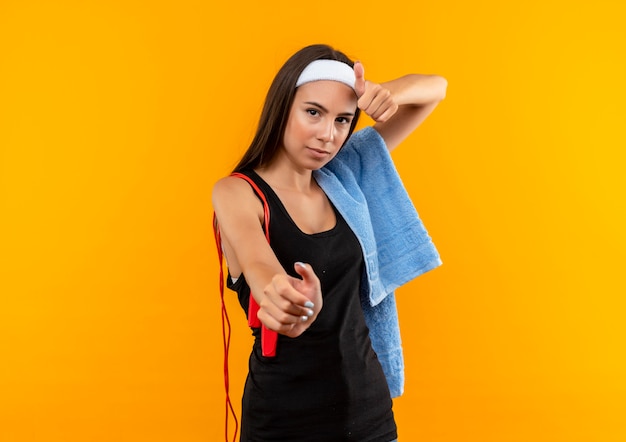 Confident young pretty sporty girl wearing headband and wristband with towel and jumping rope on her shoulders stretching out hand and showing thumb up on orange wall