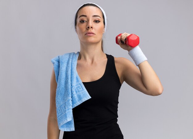 Confident young pretty sporty woman wearing headband and wristbands holding dumbbell with towel on shoulder looking at front isolated on white wall with copy space
