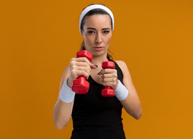 Confident young pretty sporty woman wearing headband and wristbands holding dumbbells doing boxing gesture