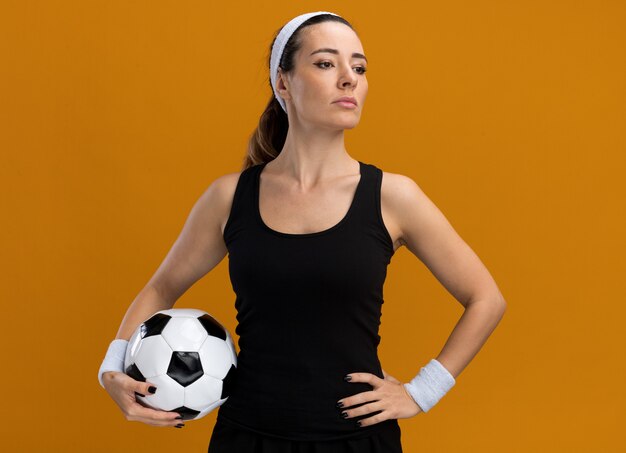 Confident young pretty sporty woman wearing headband and wristbands holding soccer ball keeping hand on waist looking at side