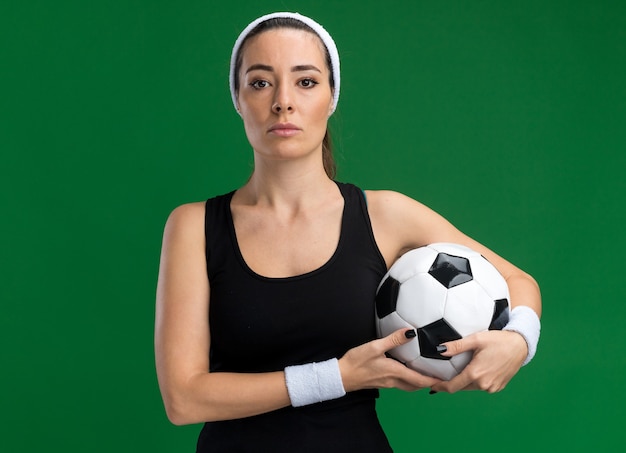 コピースペースと緑の壁で隔離の正面を見てサッカーボールを保持しているヘッドバンドとリストバンドを身に着けている自信を持って若いかなりスポーティーな女性